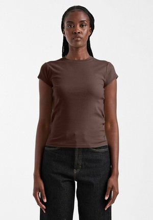 Femme aux cheveux tressés debout, portant un t-shirt marron ajusté à manches courtes et un jean foncé, sur un fond clair uni.