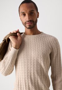 Άνδρας που φοράει μπεζ πουλόβερ με καναλέ σχέδιο, κρατώντας καφέ δερμάτινη τσάντα στον ώμο, κοιτώντας μπροστά σε απλή φόντο.