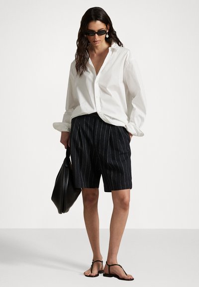 Chemise blanche à manches retroussées, associée à un short à rayures pinstripes foncé. Des sandales noires et un grand sac fourre-tout noir complètent l'ensemble.