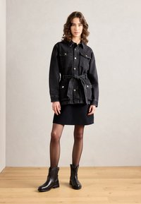 Jachetă din denim negru cu curea tip tie și buzunare pe piept, asortată cu o fustă neagră și cizme din piele până la glezne. Fundal din lemn.