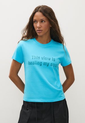 Femme aux longs cheveux ondulés portant un t-shirt bleu vif avec le texte "This view is healing my soul" et un pantalon noir, debout devant un fond uni.