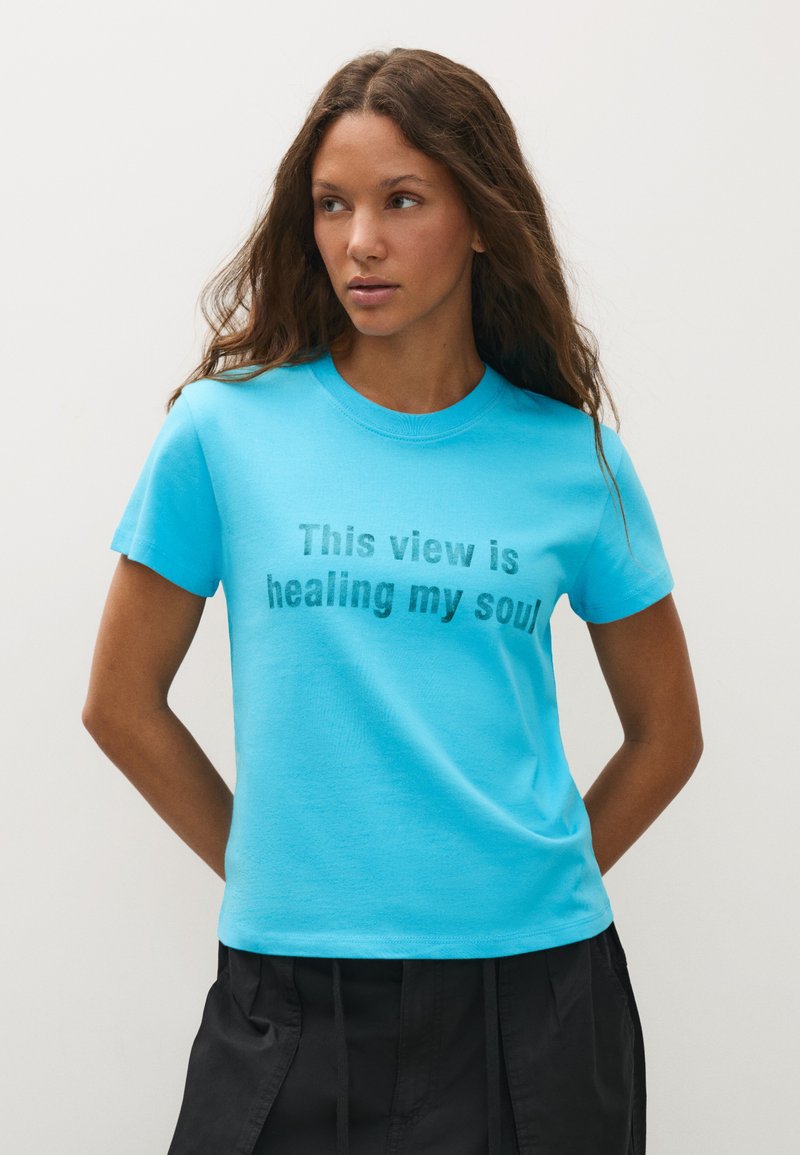 Femme aux longs cheveux ondulés portant un t-shirt bleu vif avec le texte "This view is healing my soul" et un pantalon noir, debout devant un fond uni.