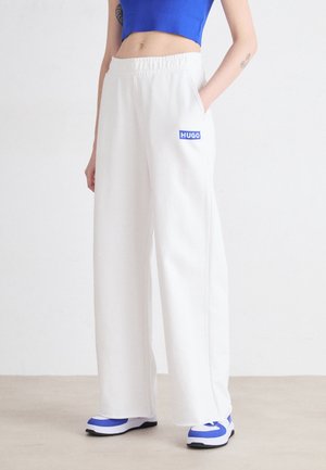 Person trägt weiße, weit geschnittene Hosen mit "HUGO"-Logo, blaues Crop-Top und weiße sowie blaue Turnschuhe, steht mit einer Hand in der Tasche.