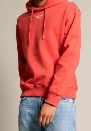 Osoba nosząca koralową bluzę z kapturem Calvin Klein Jeans oraz jasno-niebieskie dżinsy, z widocznymi logo marki na piersi i rękawie.