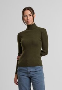 Grüner gerippter Rollkragenpullover mit langen Ärmeln, figurbetontem Design und strukturiertem Stoff, kombiniert mit hellblauen Hosen.