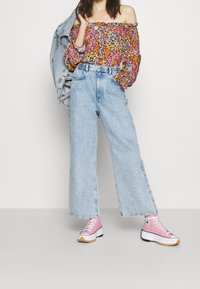 Bloemige off-shoulder top, lichte denim wijde pijpen jeans en roze high-top sneakers met witte zolen. Jas over de schouder gedrapeerd.