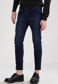 Dark blue denim jeans, slim fit, mid-rise design with a smooth texture and minimal stitching. Worn with black leather shoes.