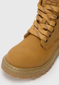 Botte imperméable beige avec bout rond, tige texturée et semelle en caoutchouc durable. Elle présente des lacets multicolores en tresses et des accents de couture.