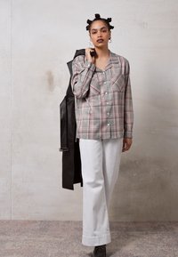 Camisa xadrez cinza, vermelha e branca com colarinho e dois bolsos no peito, combinada com calças largas brancas e uma jaqueta preta. Sapatos pretos em destaque.