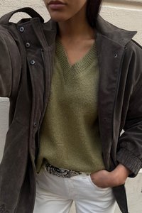 Rudas suedinė striukė su užtrauktukais ir mygtukais, žalias V formos megztinis, baltos džinsai ir raštuotas diržas su blizgiu sagtimi.