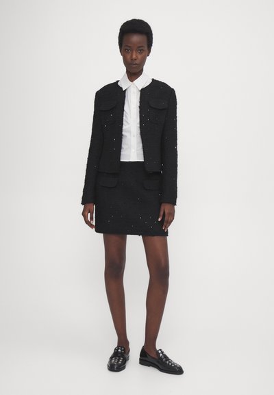 Veste en tweed noir avec poches, associée à une jupe noire texturée. Chemise à col blanc en dessous. Chaussures noires à enfiler avec accents métalliques.