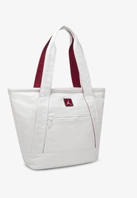 Άσπρη τσάντα καμβά με κόκκινες λεπτομέρειες, που διαθέτει μια μπροστινή τσέπη με φερμουάρ, δύο ανθεκτικούς ιμάντες και ένα λογότυπο στην μπροστινή πλευρά.