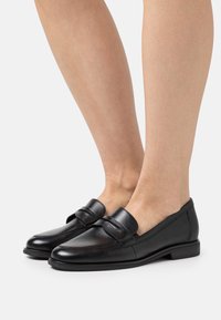 Mocasines de cuero negro con un acabado liso, que presentan una punta redondeada, una correa decorativa y detalles de costura sutiles. Tacón bajo y plano.