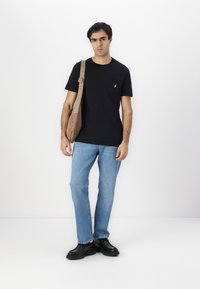 Camiseta de algodón negra con un bolsillo, combinada con jeans de color azul claro. El modelo lleva un bolso cruzado color tostado y calza unos zapatos negros.