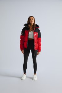 Rød og sort puffjakke med hætte, quiltet tekstur, med et logo-patch på ærmet, parret med sorte leggings og hvide sneakers.