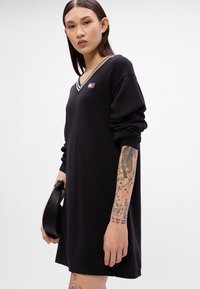 Μαύρο πλεκτό φόρεμα με μακριά μανίκια, V-λαιμόκοψη, αντίθετη ριγέ κολάρο και μικρή λεπτομέρεια λογότυπου. Το μοντέλο κρατά μια μαύρη τσάντα χεριού.