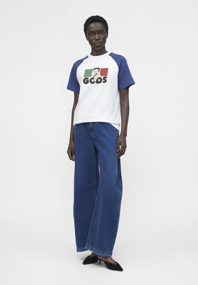 Άτομο που στέκεται φορώντας λευκό και μπλε ραγκλάν μπλουζάκι με κινούμενα σχέδια και κείμενο, μπλε τζιν με φαρδιά μπατζάκια και μαύρες μυτερές γόβες σε λευκό φόντο.