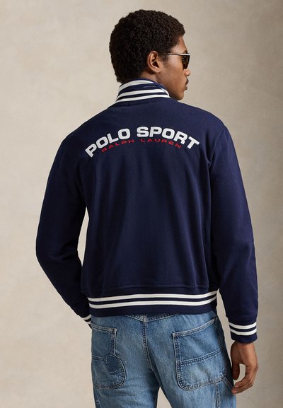 Temno modra bombažna jakna z belim in rdečim napisom "POLO SPORT" na hrbtni strani, s črtastim ovratnikom in manšetami, združena z izpranimi modrimi kavbojkami.