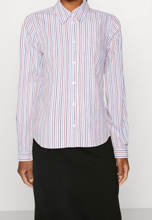 Chemise en coton à boutons avec des rayures verticales bleues et rouges, un col pointu, des manches longues et une coupe droite, portée avec une jupe noire.