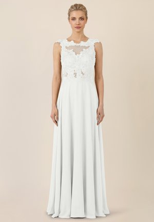 Frau trägt ein ärmelloses weißes Spitzenkleid mit floralen Mustern und einem fließenden Rock, stehend vor einem schlichten beigen Hintergrund.