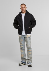 Juodas zip-up zidras su kengūros kišene, uždėtas ant baltos marškinėlių. Šviesiai mėlynos dėvėtos džinsinės kelnės su dideliais plyšiais, užsidėtos su juodai baltais sportbačiais.