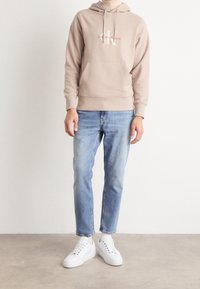 Sweat à capuche en coton beige avec une poche avant et un cordon de serrage à la capuche, orné d'un logo. Porté avec un jean en denim bleu clair et des baskets blanches.