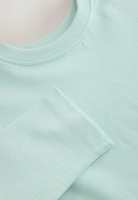 Camiseta verde menta claro hecha de algodón suave, con cuello redondo y un pequeño bolsillo triangular en el lado izquierdo.