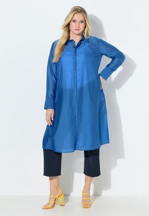 Blonde Frau steht mit den Händen in der Hüfte, trägt ein langes blaues Hemd mit Knöpfen, kurze schwarze Hosen und gelbe Sandalen mit Blockabsatz und Riemchen.