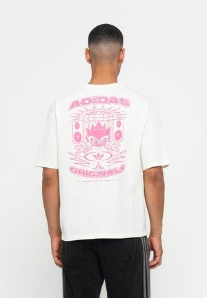 Mann med hvit Adidas Originals T-skjorte med stort rosa grafikk av en karakter, høyttalere og globe på baksiden, sammen med svarte bukser.