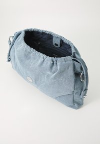 Džínová kabelka na plochu v světle modré barvě, s měkkou texturou, nařaseným vrškem a stříbrnými detaily. Interiér obsahuje podšívku s logem a zipovou kapsu.