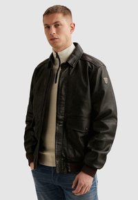 Junger Mann mit kurzen Haaren, der eine dunkelbraune Lederjacke über einem beigen Pullover mit Reißverschluss bis zur Brust und blauen Jeans trägt, mit den Händen in den Taschen stehend.