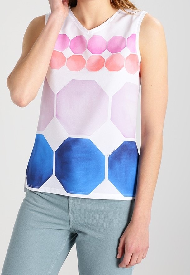Femme portant un haut blanc sans manches avec des motifs géométriques hexagonaux roses, violets et bleus, associé à un pantalon bleu clair.