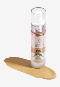 Base de maquillaje de larga duración Revolution IRL en el tono F9.5. Viene en un frasco de bomba transparente con acentos en rosa y dorado, apoyado sobre una muestra suave en beige.