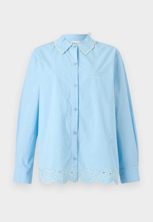 Femme debout portant une chemise bleu clair à boutons et un short assorti avec un bord en dentelle festonnée, assortis à des baskets beiges sur fond blanc.