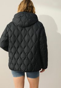 Giacca puffer nera con cappuccio oversize, caratterizzata da cuciture a forma di diamante e una texture trapuntata, indossata sopra pantaloni corti di un leggero blu.