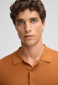 Polo shirt a maglia marrone con colletto, caratterizzato da un tessuto textured e tre bottoni alla chiusura. Vista ravvicinata della camicia.