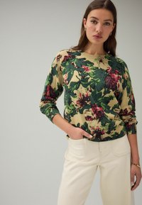 Gebreide trui met een bloemmotief in groen, bordeaux en beige. Heeft een ronde hals en lange mouwen. Gepaard met lichtgekleurde broeken.