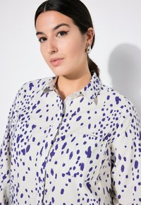 Camicia bianca con motivo a pois viola, maniche corte, colletto a punta e due tasche sul petto. Tessuto dalla consistenza liscia.