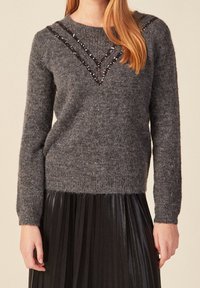 Grauer Wollpullover mit Rundhalsausschnitt, der eine V-förmige Verzierung aus schwarzen Pailletten über der Brust und gerippte Bündchen aufweist.