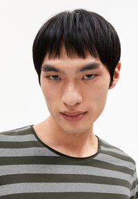 Een model met steil zwart haar draagt een groen en grijs gestreept T-shirt met ronde hals, waarbij de textuur van de stof en het kleurcontrast van dichtbij worden weergegeven.