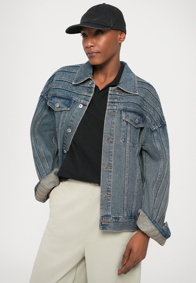 Veste en denim bleu clair avec des rayures contrastées, des boutons et des poches sur la poitrine. Portée par-dessus une chemise noire et un pantalon blanc texturé.