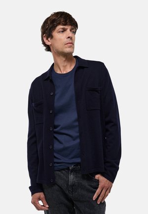 Cardigan bleu marine en tissu doux, avec une fermeture à boutons, deux poches poitrine et des manches longues, superposé sur une chemise assortie.