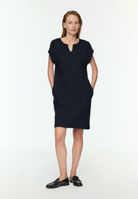 Marineblaue knielange Kleid mit Kurzärmeln, subtiler Textur, V-Ausschnitt und Seitentaschen. Kombiniert mit schwarzen Slippern.