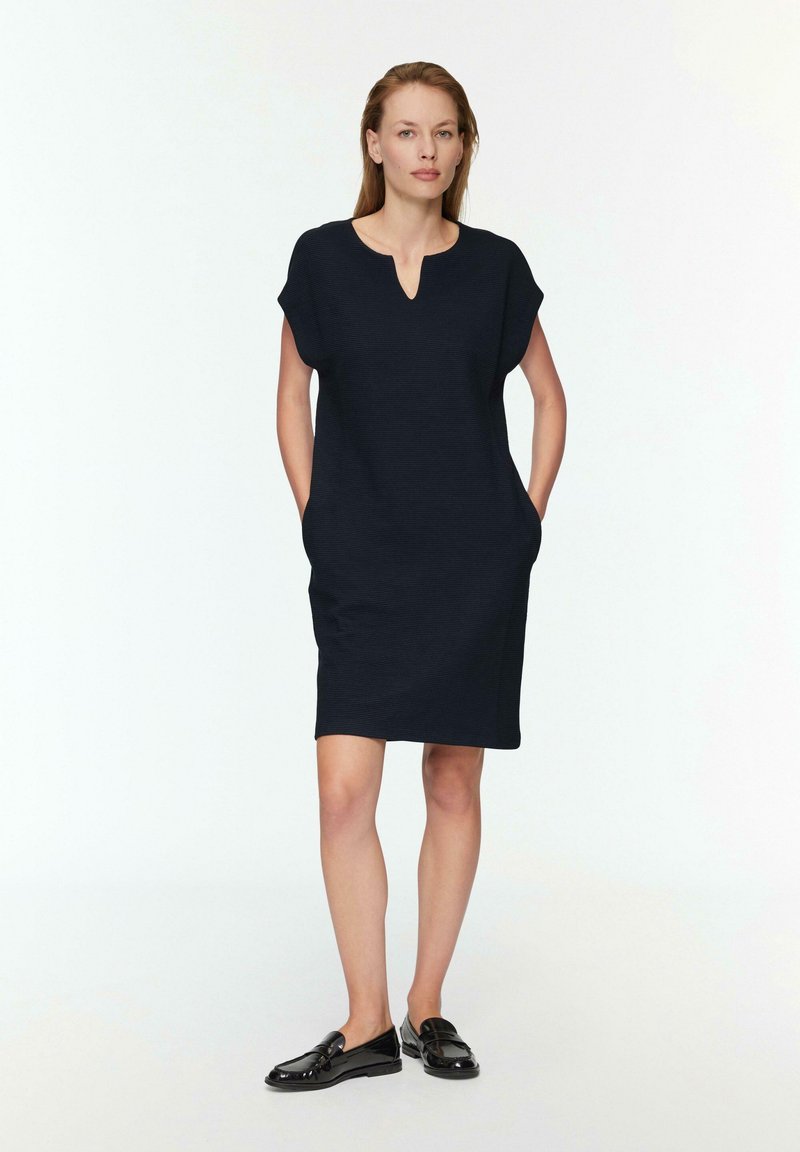 Marineblaue knielange Kleid mit Kurzärmeln, subtiler Textur, V-Ausschnitt und Seitentaschen. Kombiniert mit schwarzen Slippern.