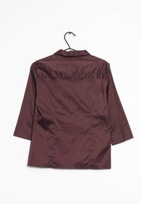 Camicia marrone a maniche lunghe con colletto, realizzata in tessuto morbido. Il retro presenta uno yoke e una sottile trama. Appesa a un gancio nero.