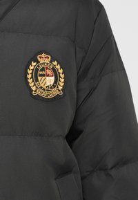 Čierna vypchávkovaná bunda s vyšívanou nášivkou erbu Ralph Lauren v zlatej a červenej farbe na hornej časti rukáva nad rozstrihnutou vreckou.