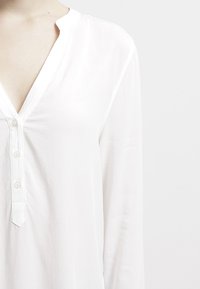 Hvid langærmet bluse med v-udskæring, knaplukning med tre knapper og glat stoftekstur. Simpelt design med rene linjer.