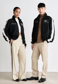 Sorte treningsjakker med hvite striper, broderte detaljer, kombinert med beige bukser og svarte sko. Modellen poserer mot en nøytral bakgrunn.