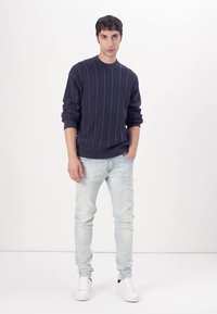 Tengerészkék, bordázott kötésű pulóver, kerek nyakkivágással, világosszürke, slim fit farmernadrággal és fehér sportcipővel viselve, egyszínű háttér előtt állva.