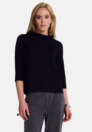 Sort langærmet top med cowl-hals og en afslappet pasform, parret med grå skræddersyede bukser. Glat stof med minimal detaljering.